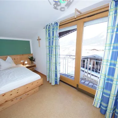 Ein gemütliches Schlafzimmer mit einem Holzbett und bunten Vorhängen. Die großen Fenster bieten einen Blick auf die umliegenden Berge.