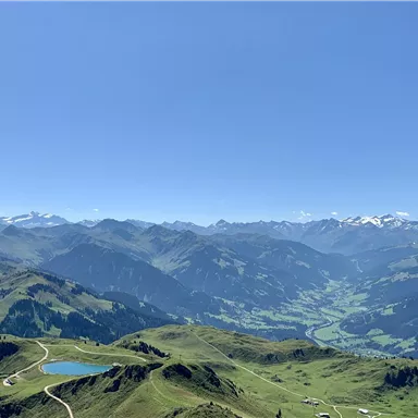 Eine atemberaubende Berglandschaft mit grünen Hügeln und schneebedeckten Gipfeln. Der klare Himmel und der strahlende Sonnenschein prägen die Aussicht.