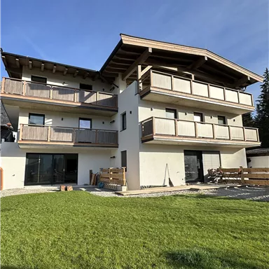 Ein modernes Haus mit mehreren Balkonen und einem grünen Garten. Der Himmel ist blau und klar, was eine einladende Atmosphäre schafft.