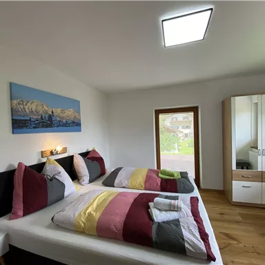 A cozy bedroom with a large bed and colorful pillows. There is a wardrobe and a view outside through a window.