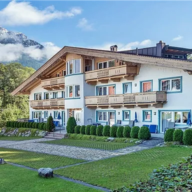 Ein malerisches Gebäude umgeben von grünen Rasenflächen und Bäumen. Im Hintergrund sind Berge und ein blauer Himmel mit Wolken zu sehen.