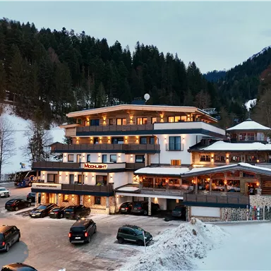 A modern hotel in the snow with surrounding mountains. Cars are parked in front of the building as the evening sky slowly darkens.