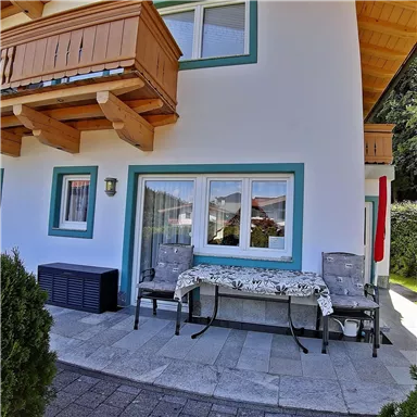 Ein gemütlicher Außenbereich mit einem Tisch und zwei Stühlen. Die Fassade des Hauses ist hell gestrichen und hat blaue Akzente.