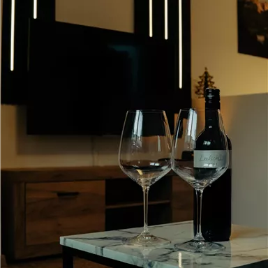 An elegant table with two wine glasses and a bottle of wine. In the background, a television and modern wall lighting can be seen.