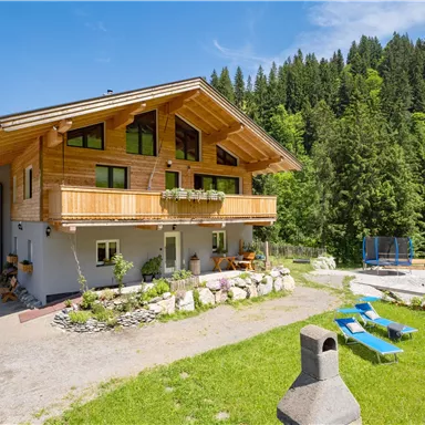 Ein schönes Holzhaus umgeben von grüner Natur. Im Vordergrund befinden sich Liegestühle und Spielgeräte für Kinder.