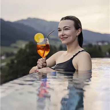 Eine Frau entspannt am Rand eines Infinity-Pools und hält ein Getränk mit einer Zitronenscheibe. Im Hintergrund sind sanfte Hügel und ein bewölkter Himmel zu sehen.