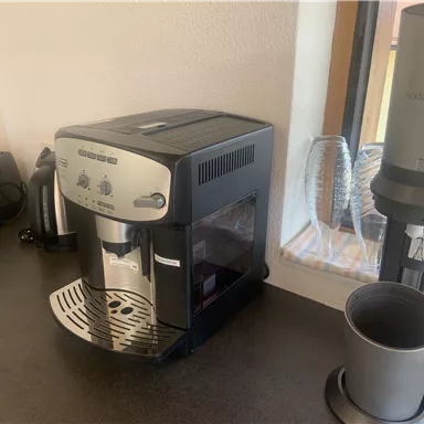 A modern coffee machine is on a countertop. In the background, a toaster and glasses are visible.