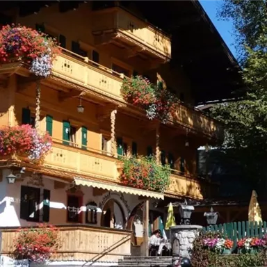 Ein traditionelles, Holzhaus mit blühenden Blumenkästen. Im Vordergrund sind Sonnenschirme und eine grüne Hecke zu sehen.