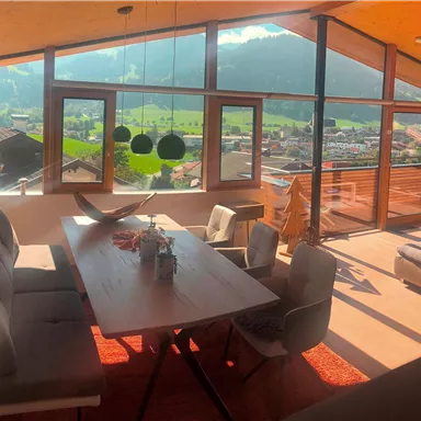 A modern living room with large windows and a view of the mountains. The space is bright and cozily furnished with a dining table and comfortable chairs.