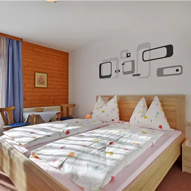 Ein gemütliches Schlafzimmer mit zwei Betten und bunten Kopfkissen. Das Zimmer hat große Fenster und eine moderne Wanddekoration.