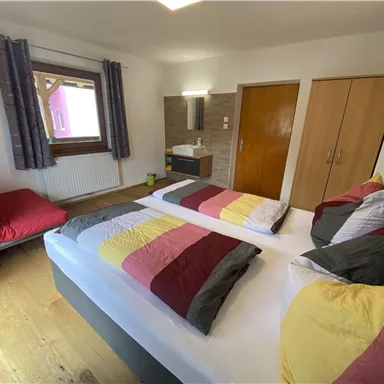 A cozy bedroom with two beds and colorful bedding. There is a desk, a chair, and a large wardrobe.