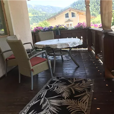 A cozy terrace with a table and chairs. In the background, there are blooming plants and a picturesque mountain landscape.