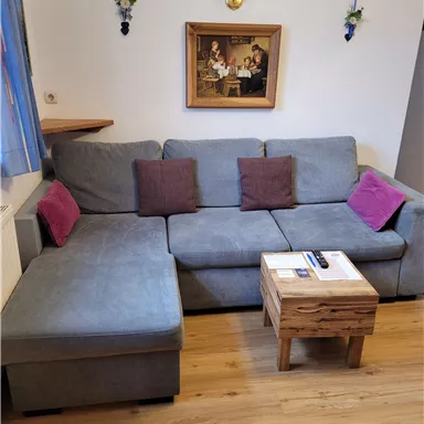 Ein gemütliches Wohnzimmer mit einem grauen Sofa und bunten Kissen. Auf einem kleinen Holztisch liegen einige Bücher und eine Fernbedienung.