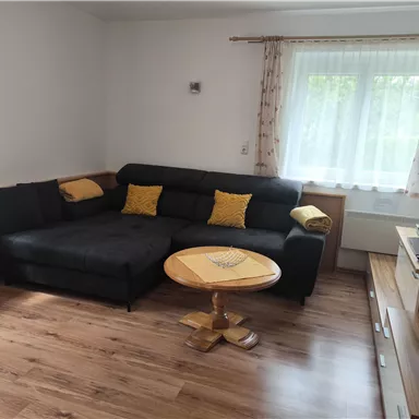 Ein gemütliches Wohnzimmer mit einer schwarzen Couch und bunten Kissen. Ein runder Tisch steht in der Mitte, und ein Fernseher ist auf einer Konsole platziert.