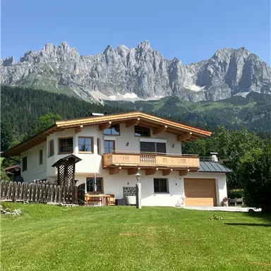 Ein wunderschönes Haus in den Alpen mit einem gepflegten Garten. Im Hintergrund ragen majestätische Berge unter klarem, blauem Himmel empor.