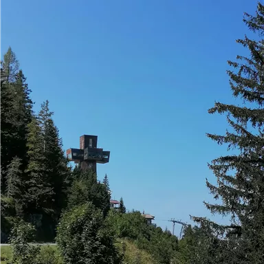 Ein großes Kreuz steht auf einem Hügel, umgeben von Bäumen. Der klare blaue Himmel sorgt für eine ruhige Stimmung.