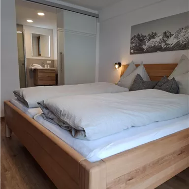 Ein modernes Schlafzimmer mit einem großen Holzbett und gemütlichen Kissen. Im Hintergrund sind ein Badezimmer und ein dekoratives Bergbild zu sehen.