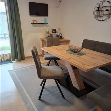 Ein gemütlicher Essbereich mit einem Holztisch und modernen Stühlen. Im Hintergrund befinden sich ein TV und eine dekorative Wandablage.