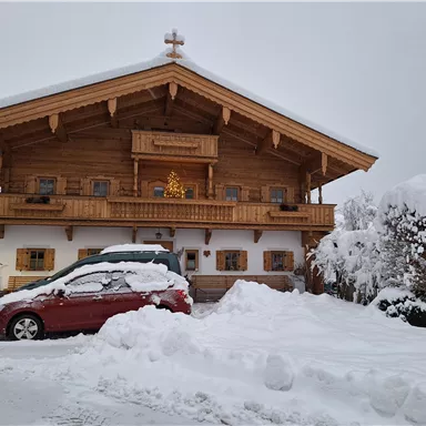 A cozy wooden house in the snow with a decorated balcony. A red car is parked in front of the house, surrounded by a blanket of snow.