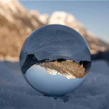 A crystal-clear ball lies on the snow and reflects the surrounding mountain landscape. In the background, snow-covered mountains and a blue sky can be seen.
