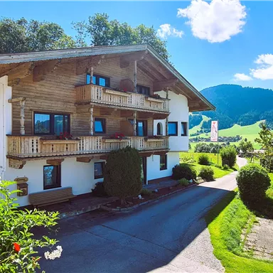 Ein charmantes alpenländisches Haus mit Holzverkleidung umgeben von grünen Wiesen und Hügeln. Der klare Himmel und die sonnige Atmosphäre schaffen eine einladende Stimmung.