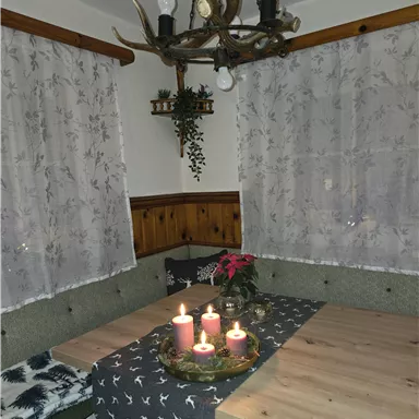 A cozy dining area with a wooden table, decorated with candles and a small plant. The windows are adorned with white curtains, and a rustic chandelier hangs from the ceiling.
