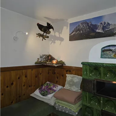 A cozy room with a wooden bench and colorful cushions. A picture of mountains hangs on the wall, while a green stove stands in the corner.