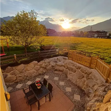 Ein idyllischer Außenbereich mit einem Tisch und Stühlen aus Holz. Im Hintergrund scheinen die Berge im Sonnenuntergang und ein blühendes Feld.