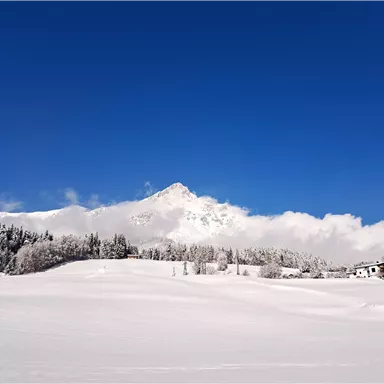 A winter landscape with snow-covered hills and an impressive mountain in the background. The sky is bright blue with a few clouds.