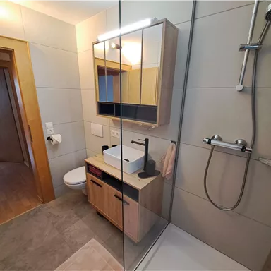 A modern bathroom with a glass shower and a simple washbasin. The walls are tiled in light shades, and there are wooden details for a warm accent.