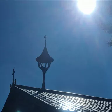 Ein sonniger Himmel über einem kleinen, traditionellen Gebäude mit einem charakteristischen Dach. Ein Wetterhahn auf dem Turm und einige Äste im Vordergrund sind sichtbar.