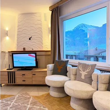 A cozy living space with modern, round armchairs and a television. The mountain landscape is clearly visible through the large window.