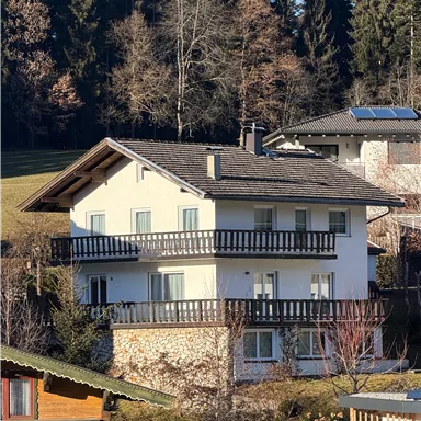 A modern house with several balconies in the midst of a green landscape. In the background, tall trees and other residential buildings are visible.