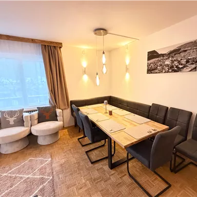A modern dining room with a large wooden table and comfortable chairs. A black and white picture hangs on the wall, and the window provides plenty of daylight.