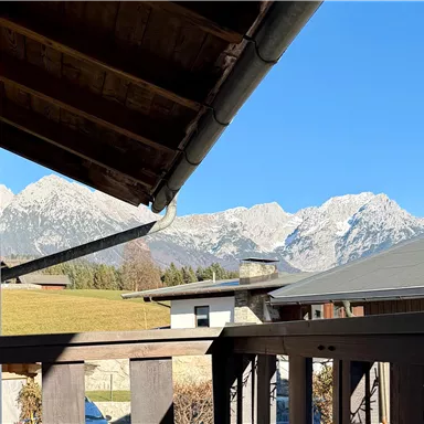 A beautiful view of snow-covered mountains under a clear blue sky. In the foreground, a balcony with a wooden railing is visible.