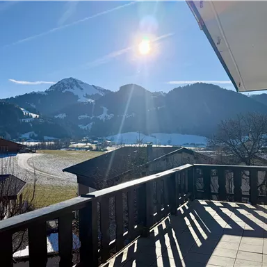 A sunny view from a balcony over green meadows and snow-covered mountains. The landscape is calm and picturesque, ideal for a relaxing day.