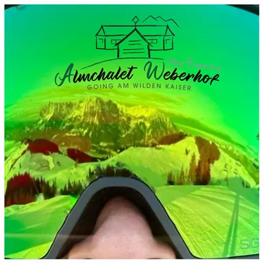Eine Skibrille mit einer grünen Linse spiegelt die schneebedeckte Berglandschaft wider. Im Hintergrund ist das Almchalets Weberhof zu sehen.