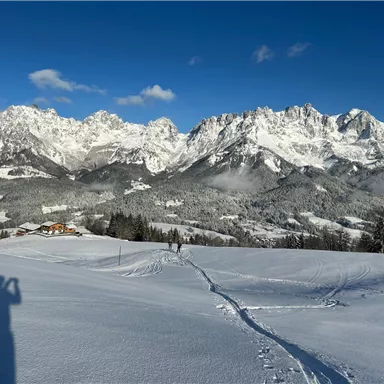 Eine winterliche Landschaft mit schneebedeckten Bergen und blauem Himmel. Im Vordergrund sind Schnee und Spuren zu sehen.