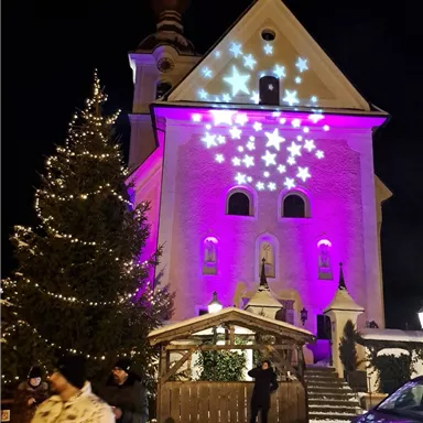 Eine festlich erleuchtete Kirche bei Nacht, umgeben von Weihnachtsbäumen. Die Fassade strahlt in lila und zeigt funkelnde Sterne.