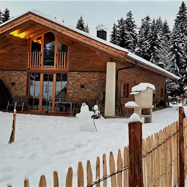 Ein modernes Holzhaus in einer verschneiten Landschaft. Im Vordergrund steht ein kleiner Schneemann und ein Zaun, umgeben von Tannenbäumen.