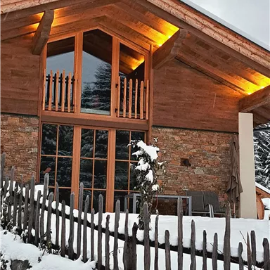 Ein gemütliches Holzhaus im Schnee mit großen Fenstern. Umgeben von einem Holzzaun und einer winterlichen Landschaft.