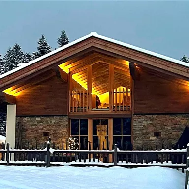 Ein modernes Chalet im Schnee mit warmem Licht. Umgeben von hohen Tannen und einer winterlichen Landschaft.