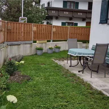 A well-maintained garden with a green lawn and colorful flowers. There is a table with chairs and a sunshade near a wall.