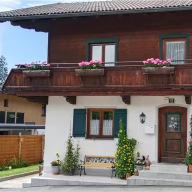 A traditional house with a wooden balcony decorated with flowers. In the foreground, there are plants and a bench.
