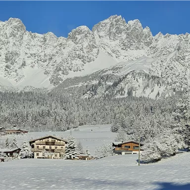 Eine verschneite Landschaft mit Bergen und einem blauen Himmel. Im Vordergrund sind einige charmante Hütten zu sehen.