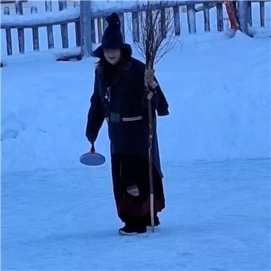 Eine Person in dunkler Kleidung steht auf einer eisbedeckten Fläche. Sie hält einen Besen und einen runden Gegenstand in der Hand.