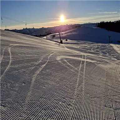 Eine verschneite Piste bei Sonnenaufgang. Der Himmel ist klar und die Spuren im Schnee sind sichtbar.