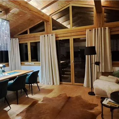A cozy living space with wooden elements and large windows. The room is modernly furnished with a dining table and a comfortable seating area.