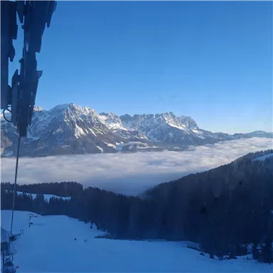 Eine beeindruckende Berglandschaft mit schneebedeckten Gipfeln und einer Wolkendecke im Tal. Der Himmel ist klar und strahlend blau.