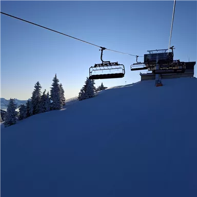 Eine schneebedeckte Landschaft mit einem Skilift, der in den blauen Himmel führt. Im Hintergrund sind winterliche Bäume zu sehen.